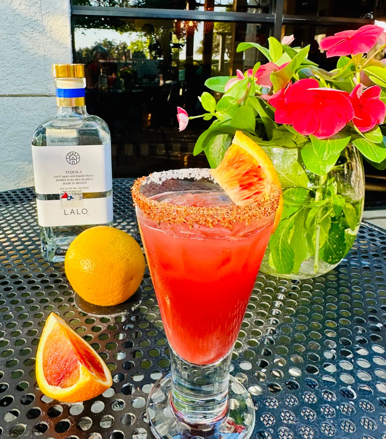 A blood-orange margarita with a chile-salt rim and a bottle of Lalo tequila on a sunny patio table at Manuel's