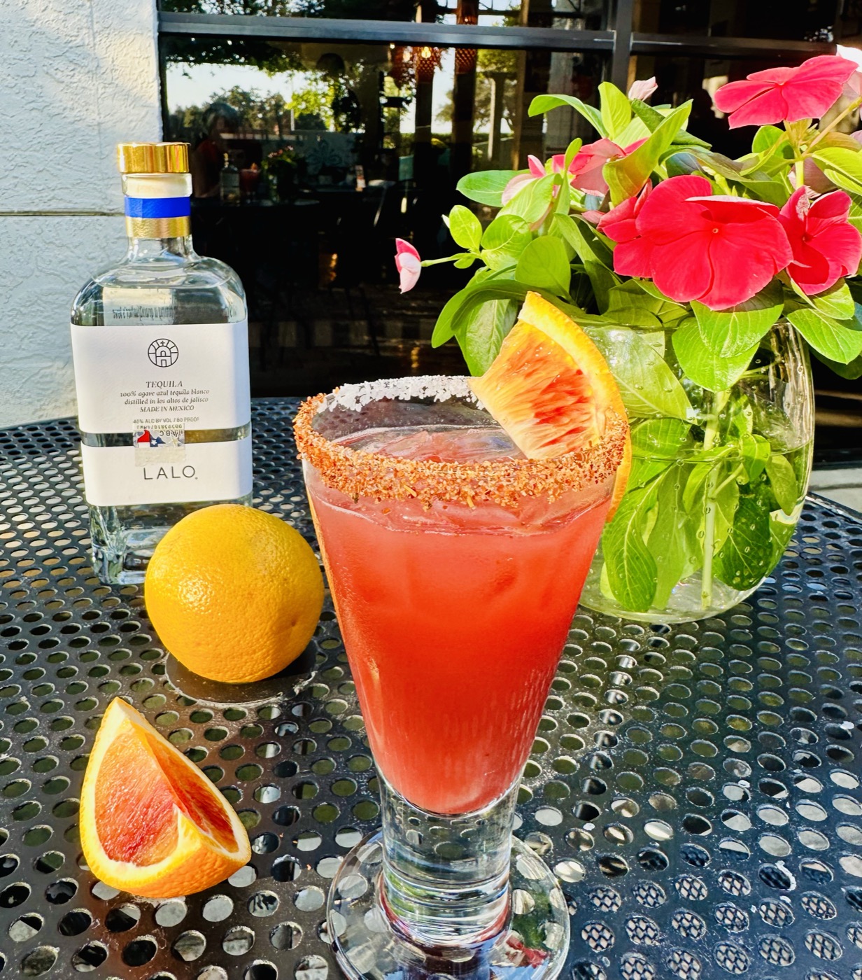 A blood-orange margarita with a chile-salt rim and a bottle of Lalo tequila on a sunny patio table at Manuel's