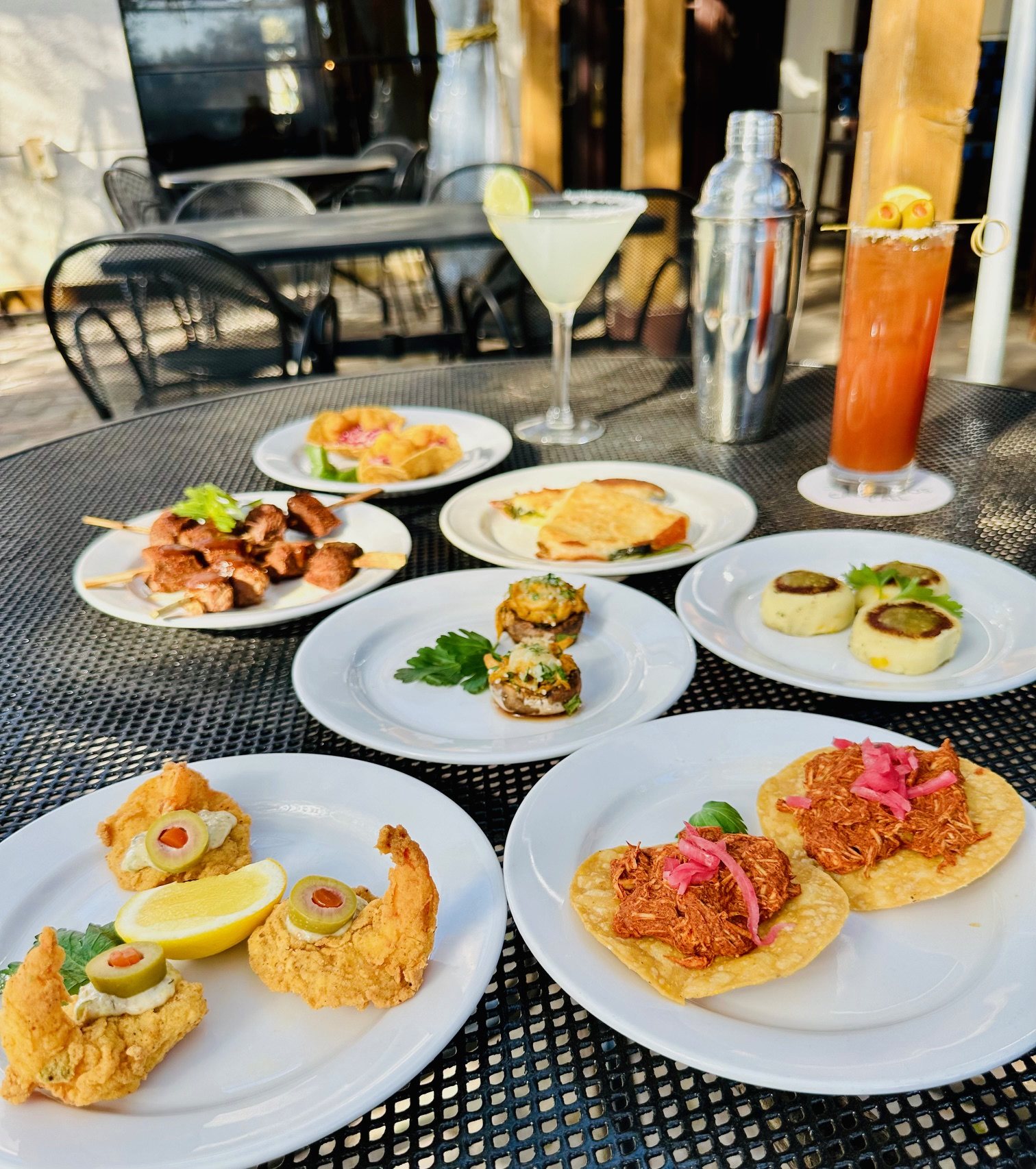 Overhead happy hour spread on Manuel's patio table — beef brochettes, chicken tostadas, crab-stuffed mushrooms, scallop sopes, scorpion shrimp, a margarita and a bloody maria